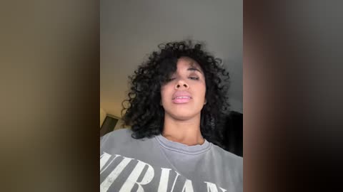 Media: Video of a Black woman with voluminous, curly black hair and light skin, wearing a gray \"Nirvana\" t-shirt, indoors with a neutral-colored wall background.