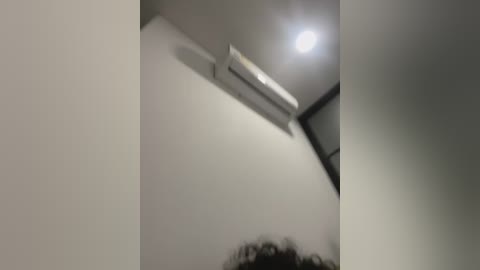 Media: A video captures a dimly lit room with a white ceiling, a large air conditioner, and a blurry person's head at the bottom. The room appears minimalistic with smooth, off-white walls.