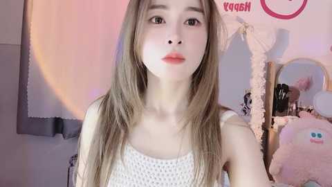 Media: Video of a young, slim, fair-skinned Asian woman with long, straight, light brown hair, wearing a white, knitted sleeveless top. She stands in a softly lit, cozy room with pink and white decor, including a plush toy and a round mirror.