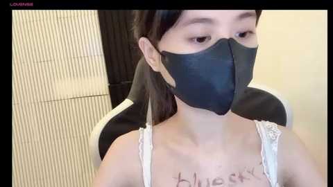 Media: Video of a young Asian woman with pale skin, short dark hair, wearing a black face mask and white lace-trimmed tank top, seated in a modern black chair, with a white tiled wall background.