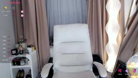 Media: Video of a modern, white ergonomic office chair facing a window with beige curtains, next to a white table holding various items, and a lamp with a wavy shade.