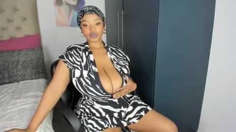 Media: Video of a Black woman with medium brown skin, wearing a low-cut zebra-patterned jumpsuit, sitting on a bed in a modern bedroom.