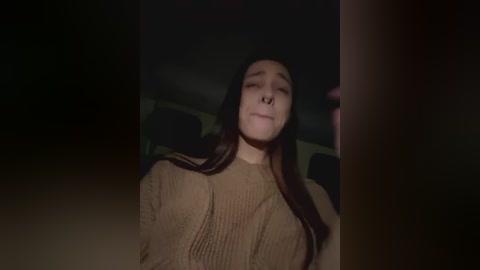 Media: Video of a young woman with long, dark hair, wearing a beige sweater, eyes closed, and a nose ring, standing in a dimly lit room, with a shadowy figure partially visible in the background.