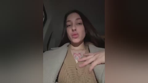 Media: A video of a young woman with long dark hair and fair skin, wearing a cream jacket and a beige knit sweater, puckering her lips in a suggestive manner.