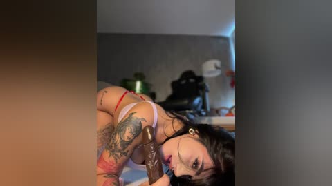 Media: A video shows a tattooed woman with dark hair, wearing red lingerie, lying on a bed in a dimly lit room. The background features a plant, a lamp, and a black statue.