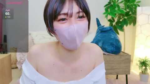 Media: A young woman with short, dark hair and pale skin wears a white off-shoulder top and a light purple face mask. She sits in a cozy bedroom with a wooden bed, a green plant, and a blue stuffed animal.