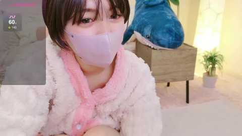 Media: Video of an Asian woman in a plush white robe with pink trim, wearing a white mask, with a blue stuffed toy and plant in the background.