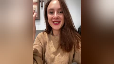 Media: Video of a smiling Caucasian woman with long, straight brown hair, wearing a beige sweater, indoors with blurred background.