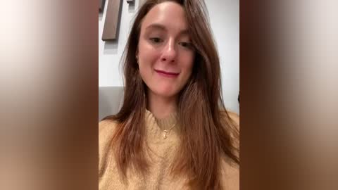 Media: Video of a young woman with long, straight brown hair, fair skin, wearing a beige knit sweater, smiling, and standing indoors with blurred background.