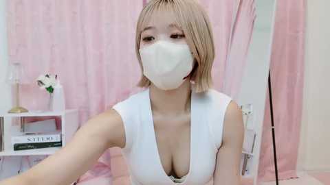 Media: Video of an Asian woman with short blonde hair, wearing a white mask and sleeveless top, sitting in a pink room with a white shelf and lamp.