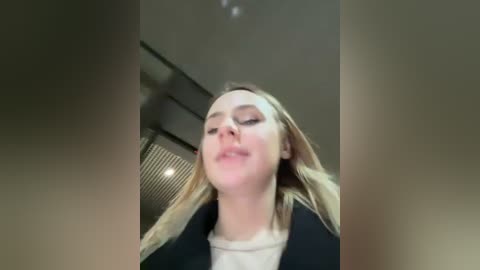 Media: Video of a blonde woman with long hair, wearing a black jacket, leaning back with eyes closed, taken from a low angle. Background shows a modern interior with a slatted ceiling and light fixtures.