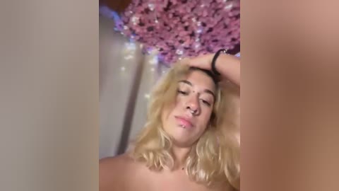 Media: Video of a blonde woman with shoulder-length hair, medium skin tone, topless, and resting her hand on her head. Background features a pink glitter curtain.