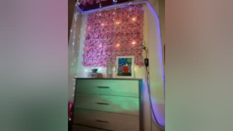 Media: Video of a cozy bedroom corner with a pink floral wallpaper backdrop, fairy lights, a white dresser, and a framed painting on the wall.