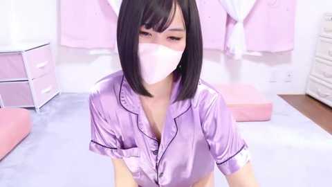 Media: Video of an Asian woman with a slim build, straight black hair, and a pale complexion, wearing a lavender satin robe and a white mask, kneeling on a pink mat in a pastel-colored room with pink and white decor.