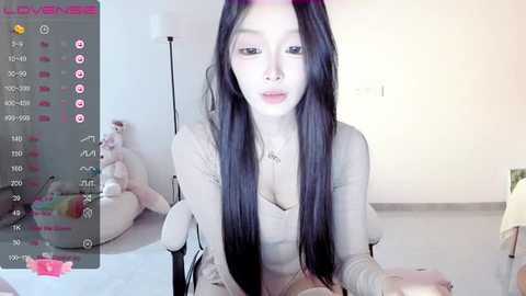 Media: Video of an East Asian woman with long black hair, pale skin, and pink lipstick, sitting in a dimly lit room, wearing a beige top, with a virtual reality headset on.