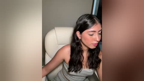 Media: Video of a young woman with long black hair, medium skin tone, wearing a gray sleeveless dress, sitting on a white leather chair, with a blurred person in the background.