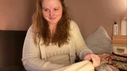 Media: Video of a smiling, fair-skinned woman with long, wavy brown hair, wearing a beige cardigan, seated on a bed with a striped pillow and patterned quilt.