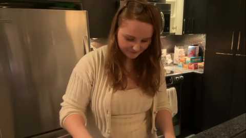 Media: Video of a Caucasian woman with long brown hair, wearing a beige cardigan over a white top, chopping vegetables in a modern kitchen with stainless steel appliances and dark cabinets.