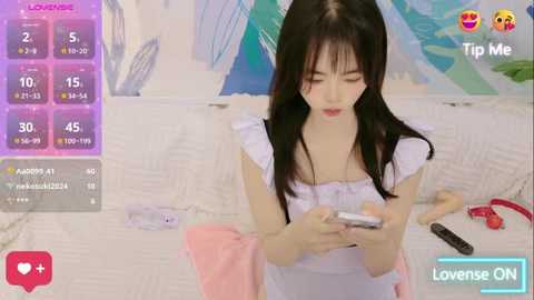 Media: Video of an Asian woman with long black hair, wearing a white dress, holding a remote control, in a white room with a bed and TV.