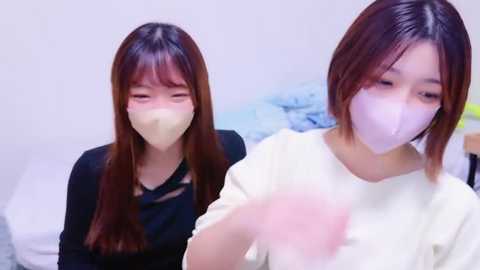 Media: Video of two young East Asian women with long, straight brown hair, wearing black tops and surgical masks, sitting on a bed with blue sheets in a minimalistic room.