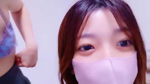 Media: Video of an Asian woman with fair skin and shoulder-length brown hair, wearing a pink face mask, looking worried. She's in a light-colored, patterned bra, and the background is plain and light.