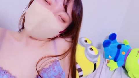 Media: Video of a young woman with straight brown hair, wearing a light blue lace bra, with a white mask covering her mouth, lying on a bed with colorful stuffed toys.