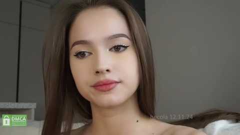 Media: A video of a young woman with fair skin, brown hair, and striking makeup, including bold winged eyeliner, pink lipstick, and subtle blush. She appears serene, gazing directly at the camera. Background features a minimalist, neutral-toned room.