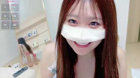 Media: A video of an Asian woman with long, straight, dark brown hair, wearing a white medical tape over her mouth, smiling. The background features a modern, white bathroom with a mirror, sink, and toiletries.