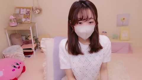 Media: A video of an East Asian woman with straight, shoulder-length brown hair, wearing a white, short-sleeved knit dress, face mask, and a pink headband with a bow. Background features a tidy, pastel-colored room with a desk, shelves, and framed art.