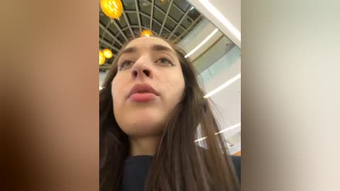 Media: Video of a young woman with long brown hair, light skin, and full lips, looking upward. Background features a modern, glass-enclosed ceiling with yellow lights.