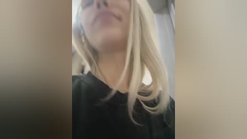 Media: Video of a woman with fair skin and blonde hair, wearing a black shirt, seen from above. She has a closed-mouth smile, and the background is blurred, suggesting an indoor setting.