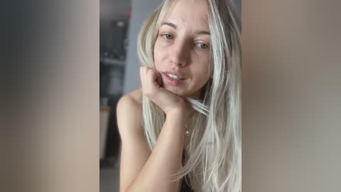 Media: Video of a young Caucasian woman with long, platinum blonde hair, fair skin, and small breasts, leaning on her hand in a contemplative pose. Background is blurred, possibly an indoor setting with muted colors.