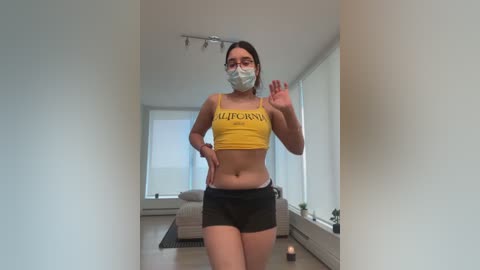 Media: Video of a young woman with light skin, medium build, wearing a yellow crop top, black shorts, and a mask, waving, in a modern, minimalistic living room.