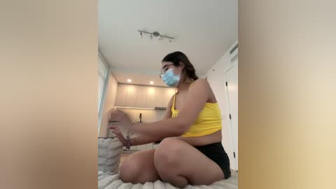 Media: Video of a young woman with medium skin tone, wearing a yellow tank top, black shorts, and a blue mask, kneeling on a white towel, in a modern, beige-toned bathroom.