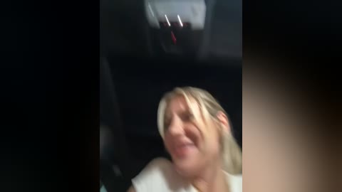 Media: Video of a blonde woman with short hair, wearing a white shirt, smiling and looking up, with a dark background, possibly inside a car.