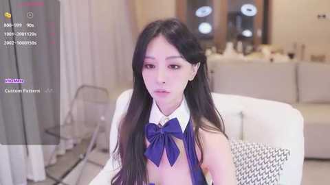 Media: Video of an Asian woman with long black hair, wearing a white blouse with a blue bow, sitting in a modern, minimalistic living room with beige walls and furniture.