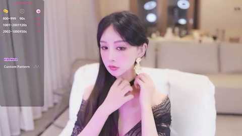 Media: A video of an East Asian woman with long black hair, wearing a black lace top, adjusting her necklace. She sits on a white couch in a modern living room with neutral tones and soft lighting.