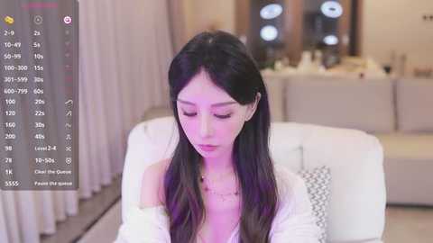 Media: Video of a young East Asian woman with long black hair, wearing a white off-shoulder top, sitting in a modern living room.