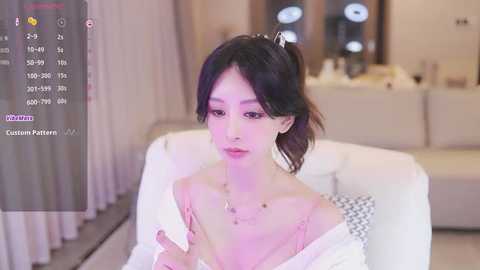 Media: A video of an East Asian woman with pale skin, black hair in a ponytail, wearing a white off-shoulder top, sitting in a modern, minimalist living room with beige walls and a white couch.