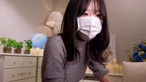 Media: Video of a young East Asian woman with long black hair, wearing a gray sweater and a white surgical mask, seated indoors with a beige dresser and potted plants in the background.