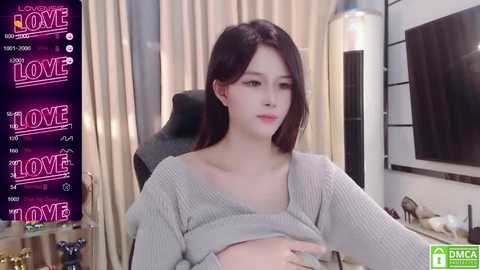 Media: Video of a young Asian woman with long, dark hair, wearing a grey knitted sweater, sitting in a chair in a modern living room.