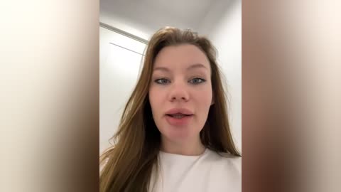 Media: Video of a fair-skinned woman with long, straight brown hair and light makeup, wearing a white shirt. She has a neutral expression and is standing in a minimalist, light-colored room with a ceiling light visible in the background.