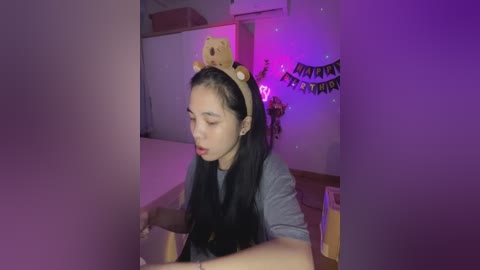 Media: Video of a young woman with long black hair, wearing a teddy bear headband, a grey shirt, and a stunned expression, in a dimly lit room with purple and yellow decorations.