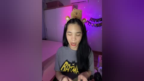 Media: Video of a young Asian woman with long black hair, wearing a gray \"Beyonc\u00e9\" t-shirt and a teddy bear headband, indoors with purple lighting, holding a phone.