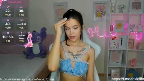 Media: Video of a young woman with light skin, long dark hair, and a small bust, wearing a blue checkered bra, sitting on a bed in a bedroom with pink neon lights, plush toys, and a framed poster.