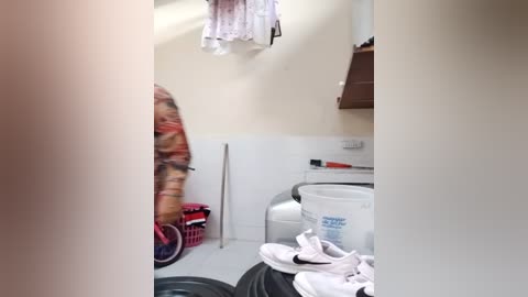 Media: Video of a small laundry room with white walls, a washer and dryer, a pink laundry basket, a mop, and a pile of white clothing on the floor.