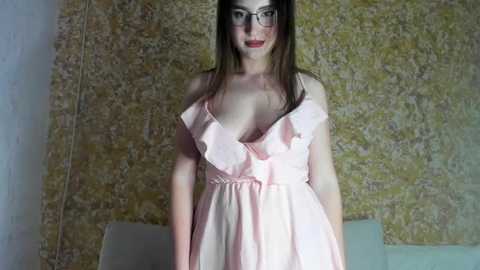 Media: A video of a fair-skinned woman with long brown hair, wearing black-rimmed glasses and a low-cut, ruffled pink dress, standing in front of a textured, yellowish-green wall.