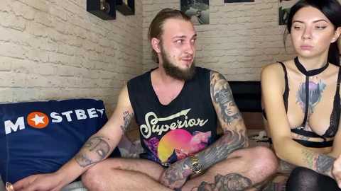 Media: Video of a tattooed, bearded man in a black tank top and woman in a black choker and bra, sitting cross-legged on a bed with a brick wall background.