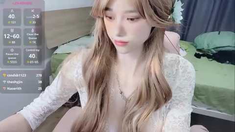 Media: Video of a young East Asian woman with long, wavy blonde hair, wearing a white lace cardigan and a light-colored dress, sitting on a bed in a bedroom. Live stream overlay shows viewers and chat.