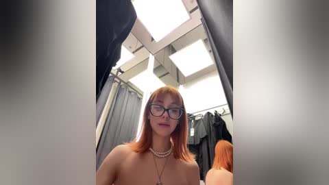 Media: Video of a topless, fair-skinned woman with red hair, wearing black-framed glasses, standing in a brightly lit changing room.